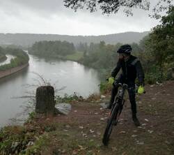 Radfahrer mit Helm im Regen auf einem Weg oberhalb eines Flusses