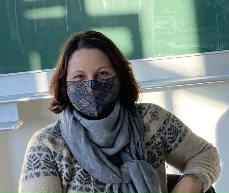 Frau mit FFP2-Maske und Schal sitzt in einem Klassenraum vor einer grünen Tafel