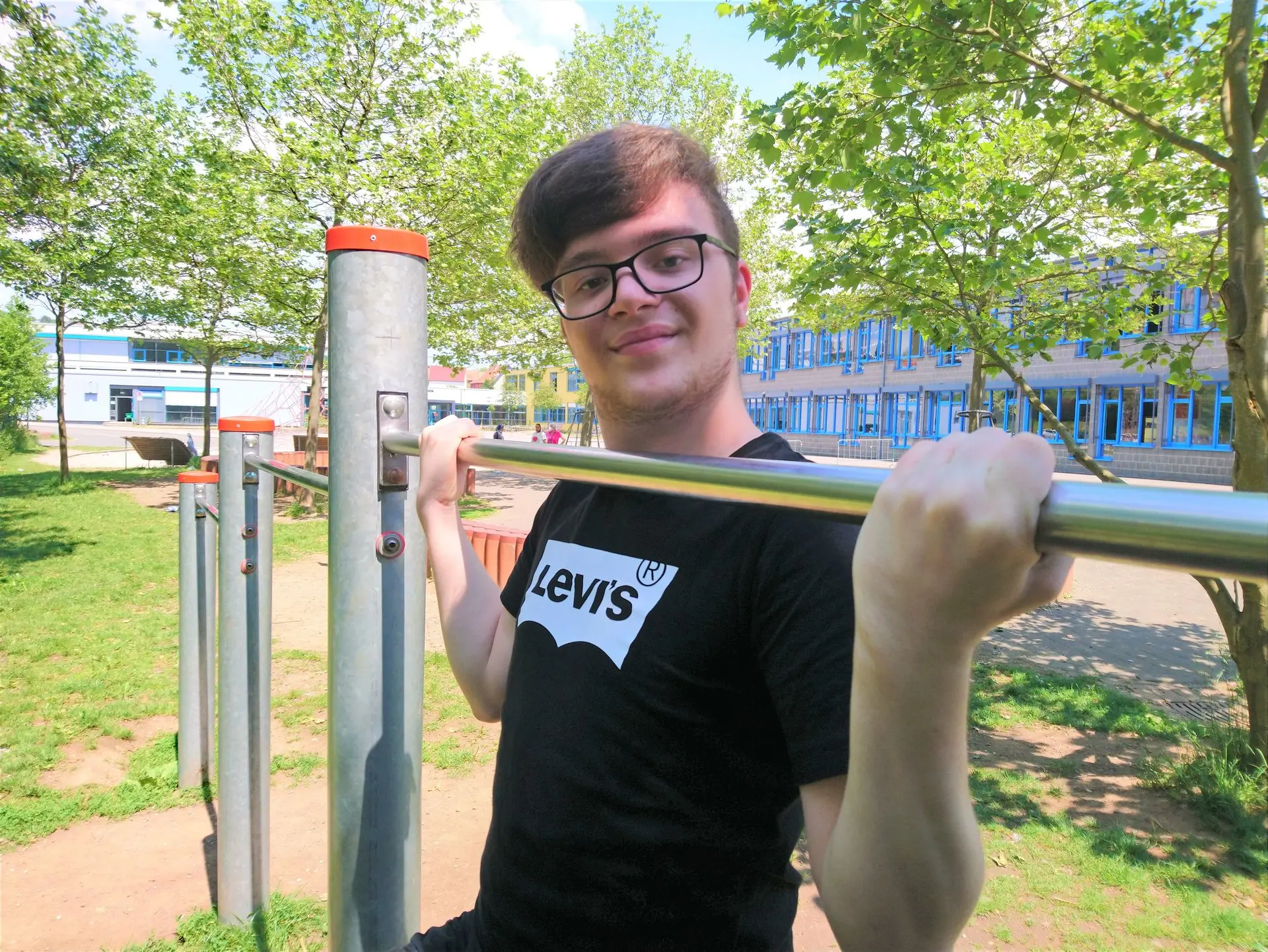 Junger Mann mit Brille hängt lächelnd an einer Reckstange auf einer Outdoor-Fitnessanlage