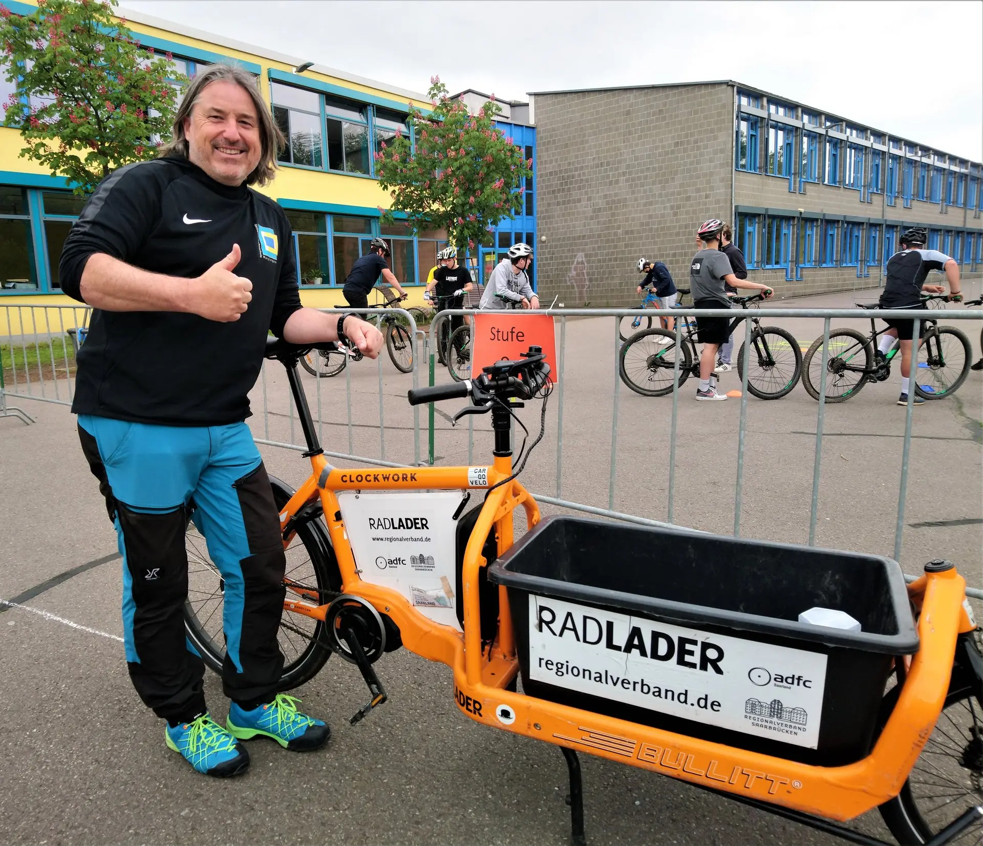 Mann steht neben einem orangefarbenen Lastenrad mit Aufschrift „Radlader“ auf dem Schulhof