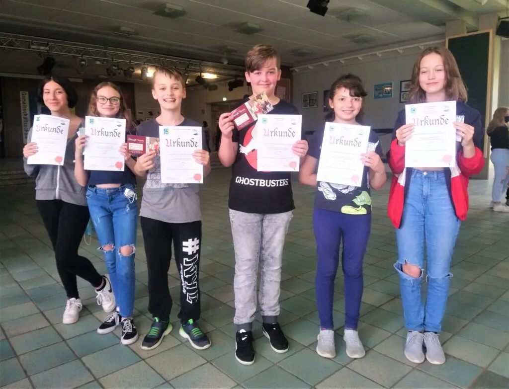 Fünf Schülerinnen und Schüler stehen in der Aula und halten Urkunden hoch