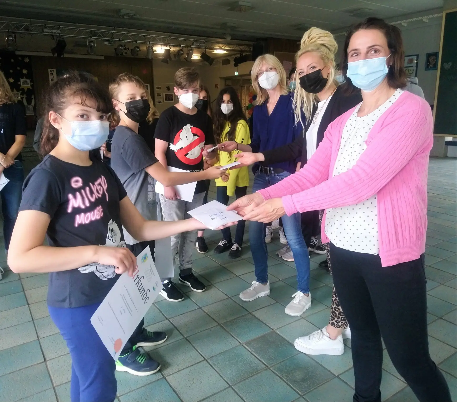 Lehrerin mit Maske überreicht einer Schülerin eine Urkunde, weitere Jugendliche schauen zu