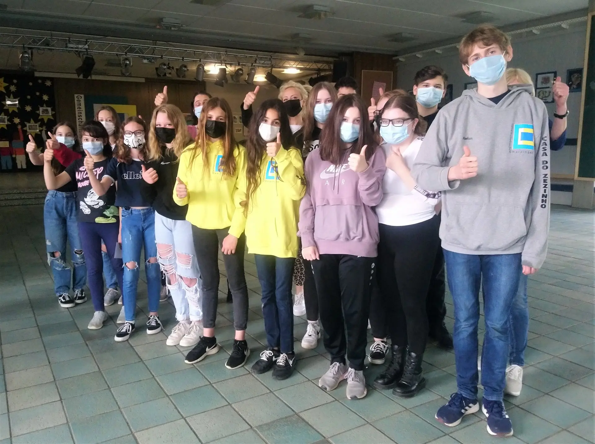 Gruppe von Schülerinnen und Schülern mit Masken posiert in der Aula, einige zeigen Daumen hoch