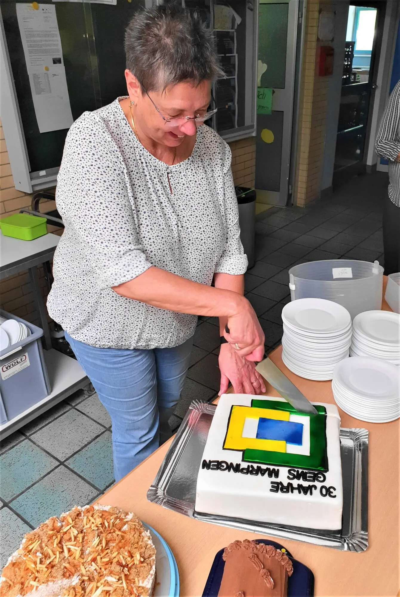 Frau mit grauem Pullover schneidet bei einer Feier eine Torte mit dem Schullogo an