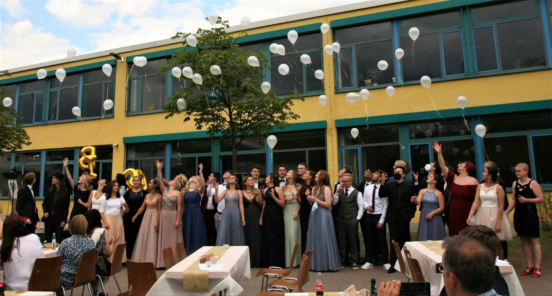 Festlich gekleidete Abiturientinnen und Abiturienten auf dem Schulhof vor dem Gebäude mit Luftballons