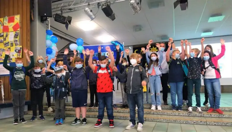 Schülergruppe jubelt mit erhobenen Armen auf der Bühne der Aula, dekoriert mit Luftballons