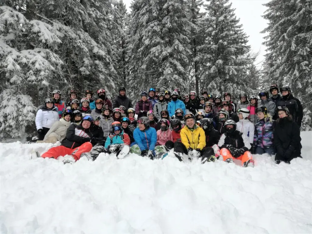Große Schülergruppe sitzt und steht im Schnee vor verschneiten Tannen