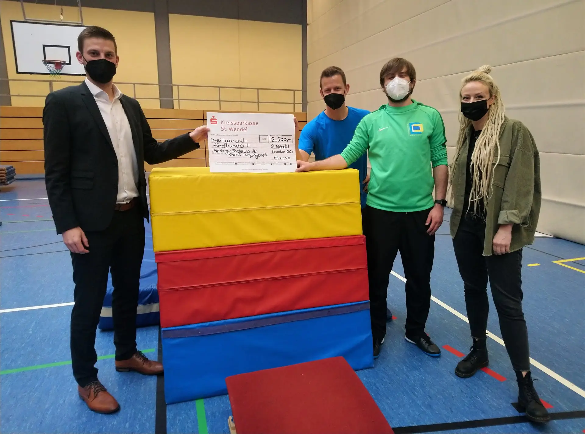 Zwei Männer und eine Frau mit Masken bei der Übergabe eines bunten Turnkastens in der Sporthalle