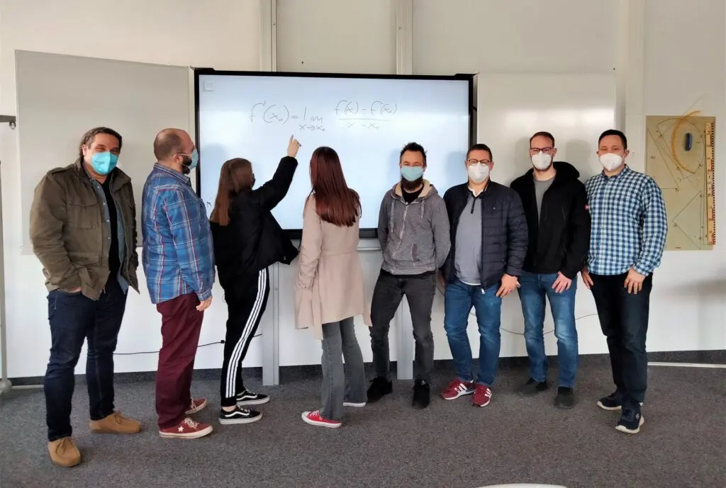Gruppe Erwachsener mit Masken steht vor einem neuen digitalen Whiteboard im Klassenraum