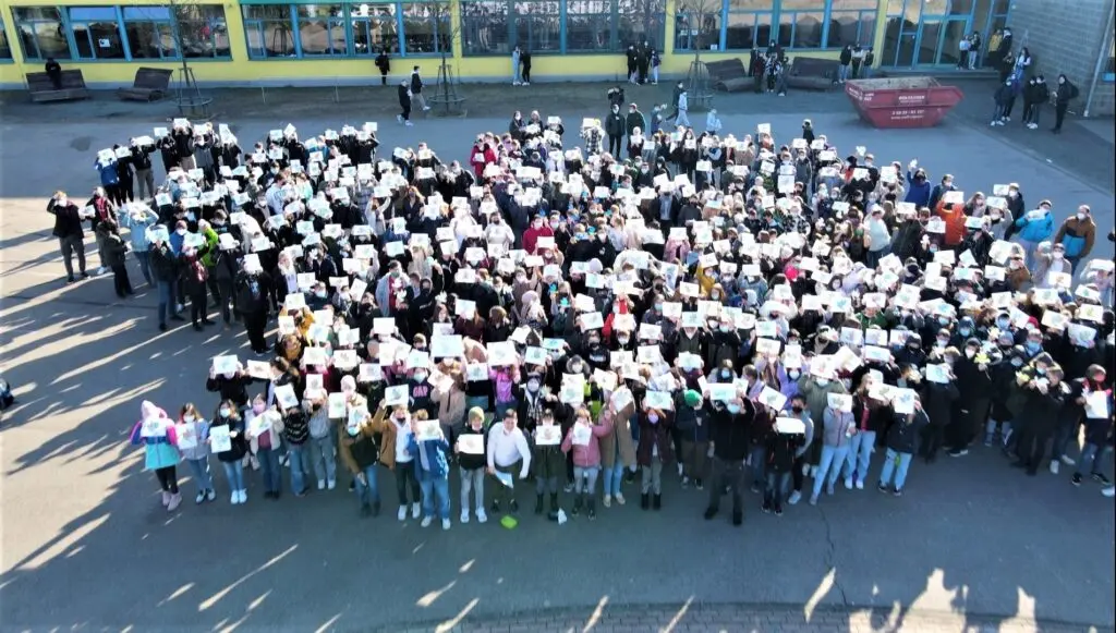 Luftaufnahme: viele Schülerinnen und Schüler bilden auf dem Schulhof eine Figur als Friedenszeichen