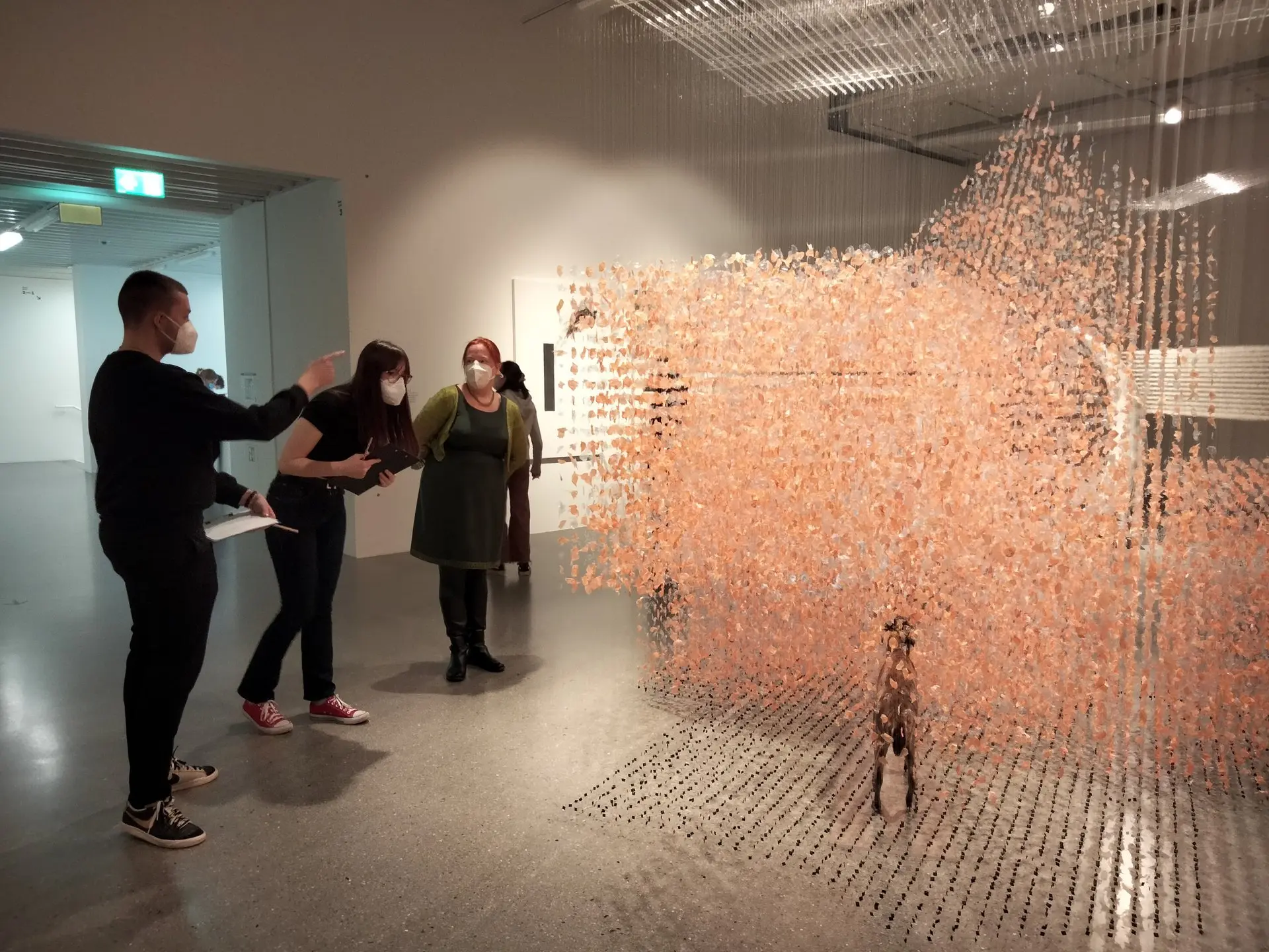 Besucher stehen in einem Museumsraum vor einer großen Installation aus vielen orangefarbenen Fäden