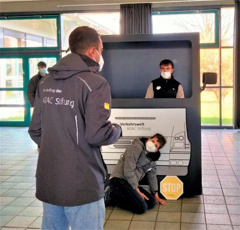 Schüler sitzt in einem Fahrsimulator in Form einer LKW-Fahrerkabine, ein Mann gibt Erklärungen