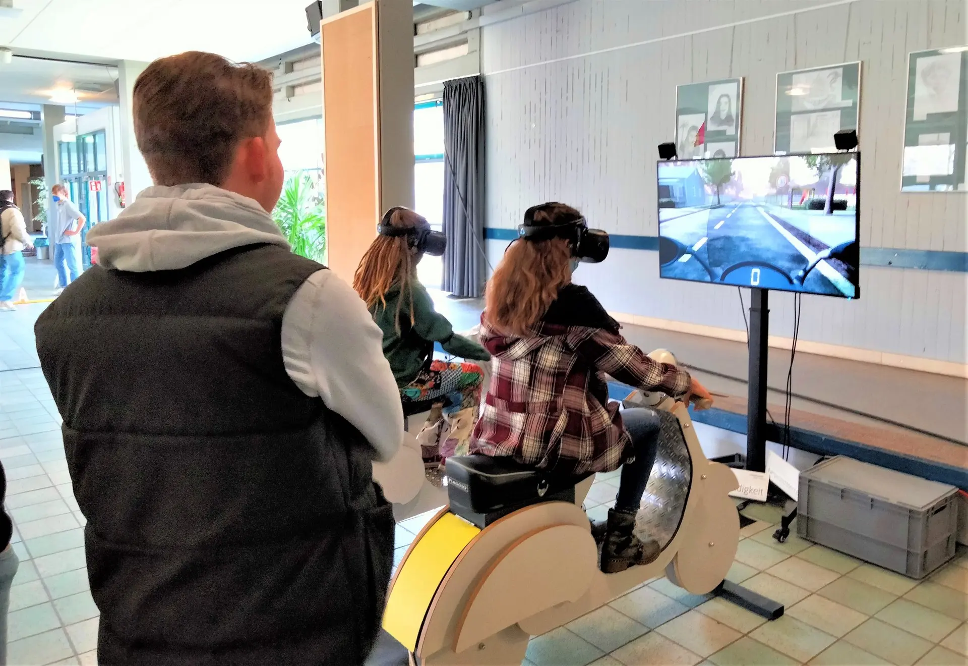 Jugendliche probiert mit VR-Brille einen Fahrsimulator aus, ein Mann schaut zu