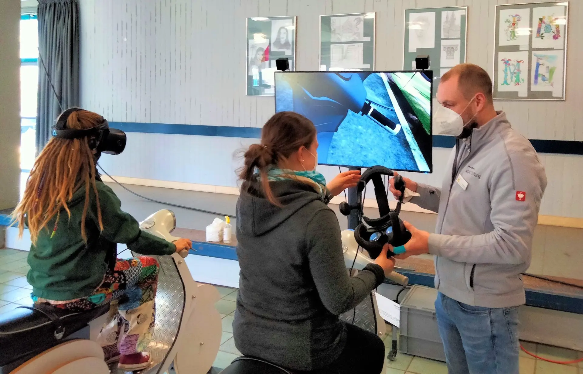 Schülerin mit VR-Brille an einem Fahrradsimulator, daneben ein Betreuer und ein Bildschirm