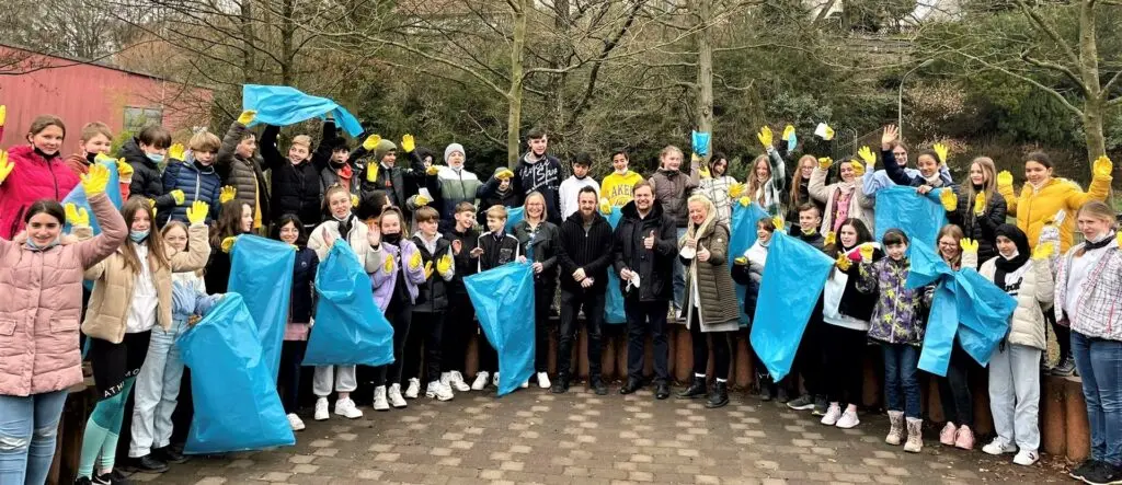 Große Schülergruppe mit hellblauen Müllsäcken und gelben Westen auf einem Schulhof
