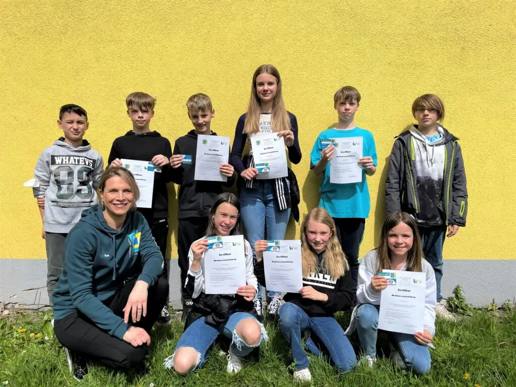 Schülergruppe mit Lehrerin zeigt Urkunden vor einer gelben Wand nach der Streitschlichter-Ausbildung