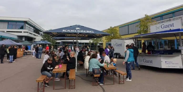 Schulhof mit Pavillons, Tischen und vielen Menschen bei einem Schulfest