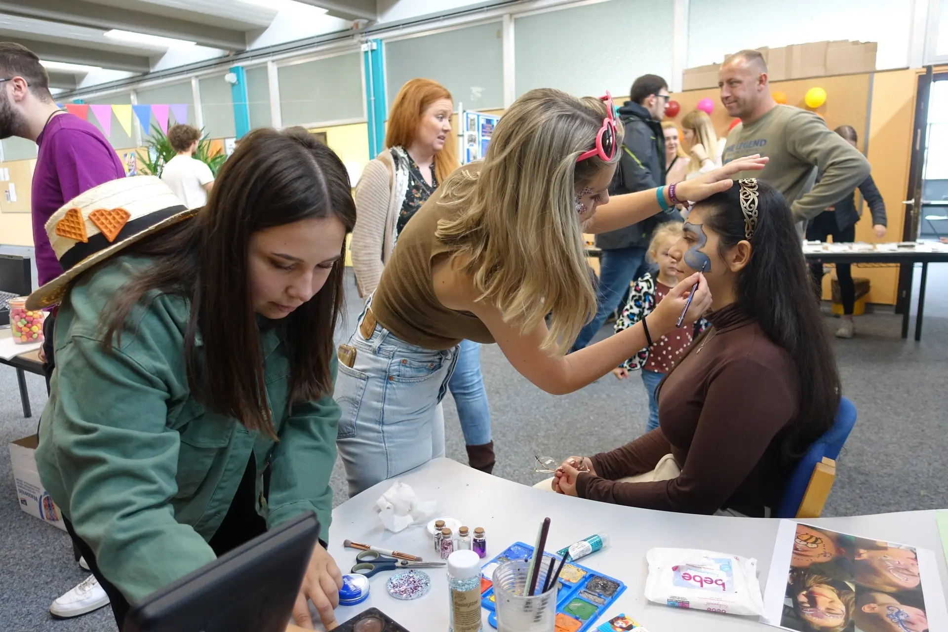 Schülerinnen schminken beim Schulfest Kinder an einem Tisch mit Schminkfarben
