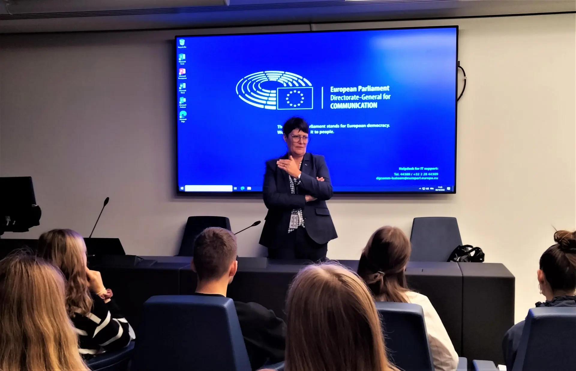 Frau spricht vor einem großen Bildschirm zu einer Schülergruppe in einem Konferenzraum