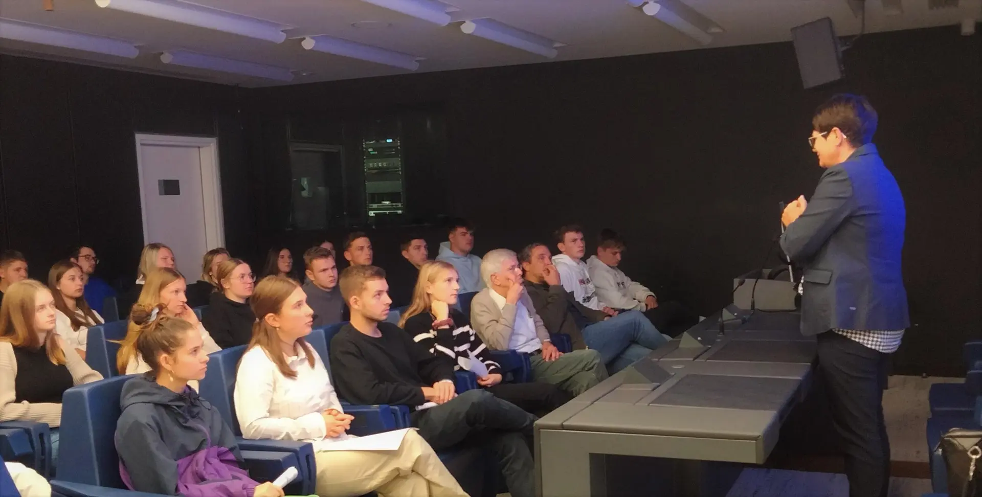 Schülergruppe sitzt in einem dunklen Konferenzraum und hört einem Redner zu