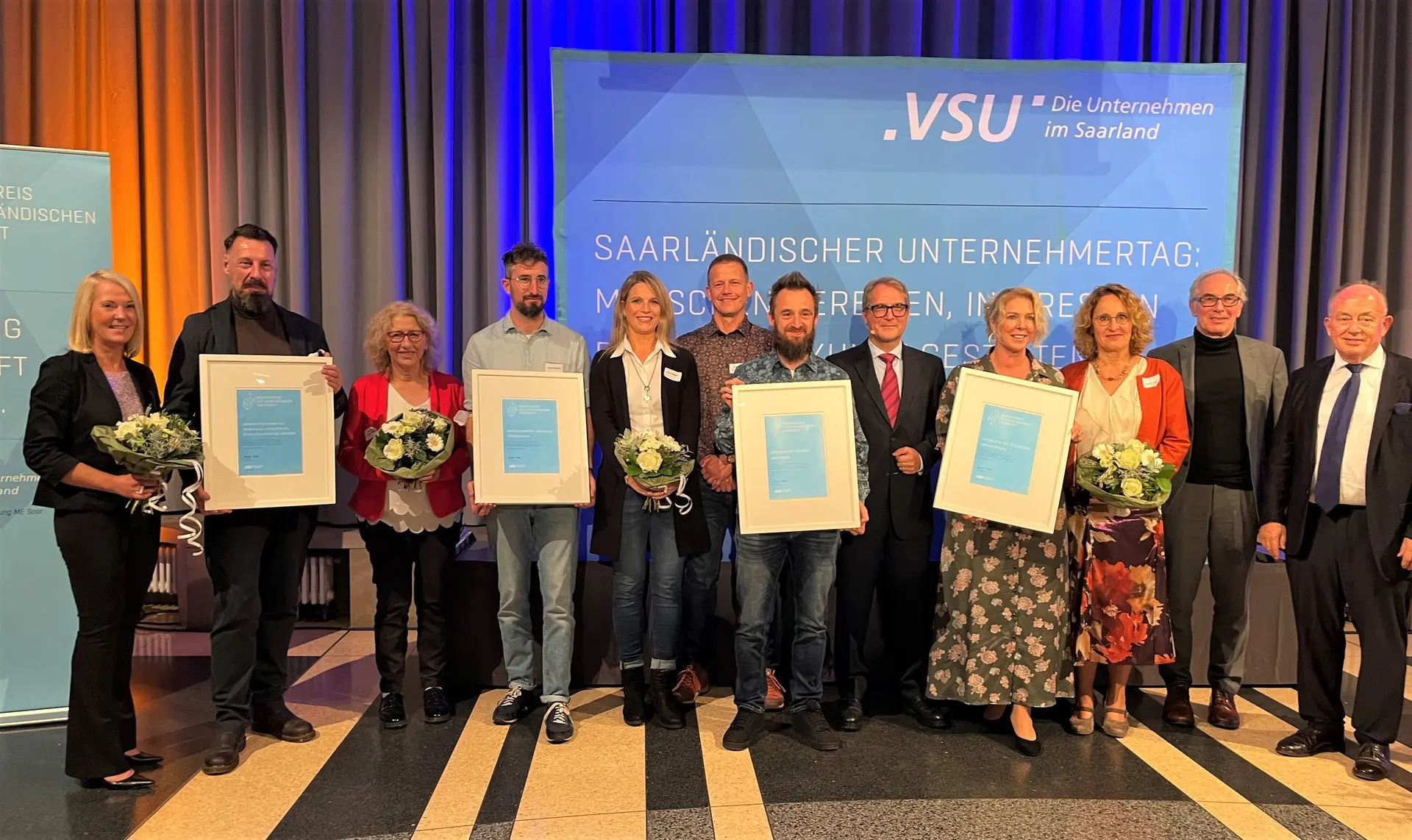 Preisträgerinnen und Preisträger mit Urkunden und Blumen auf einer Bühne vor VSU-Logo