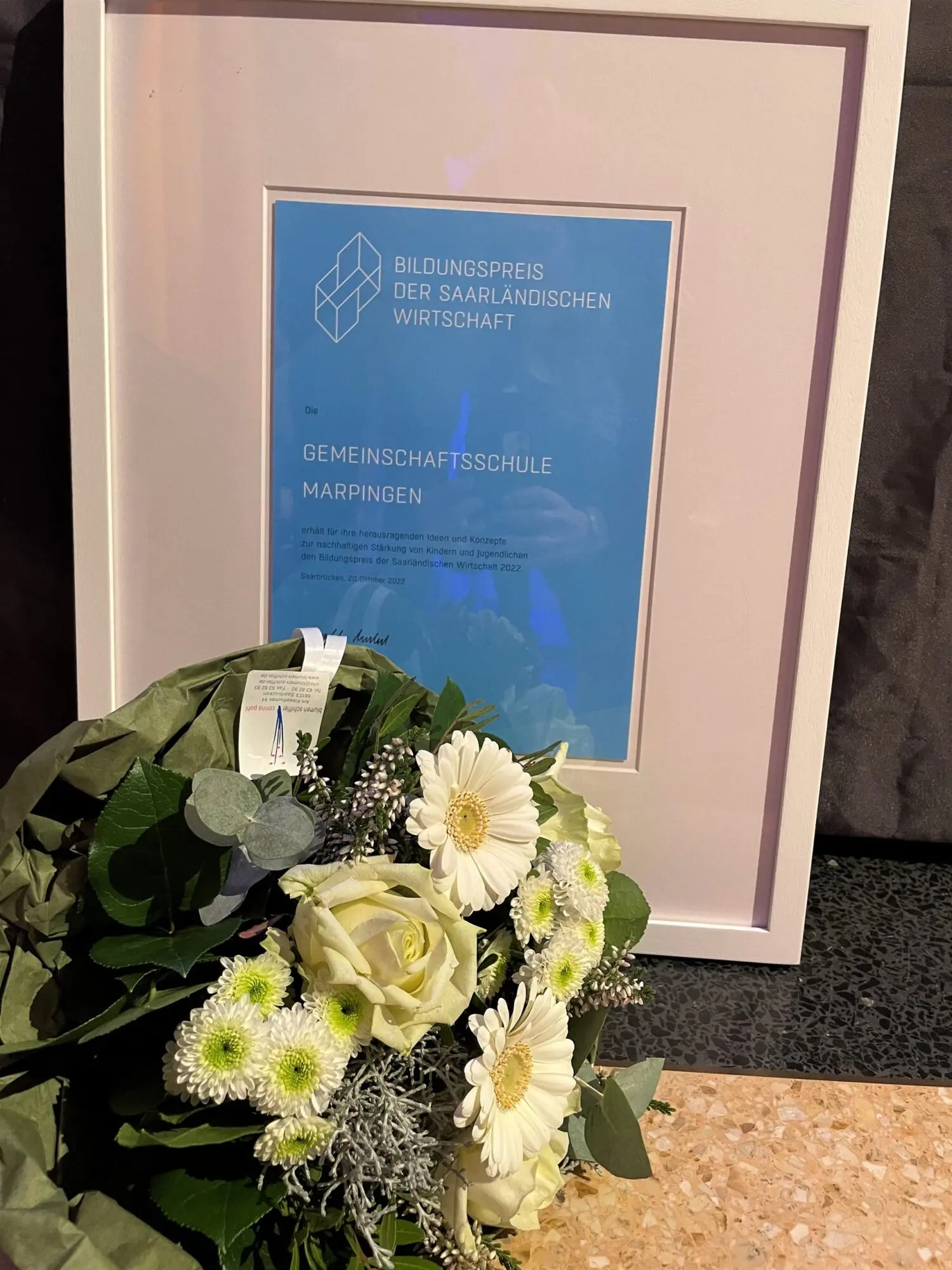 Gerahmte Urkunde, an eine Wand gelehnt, davor ein Blumenstrauß mit weißen Rosen