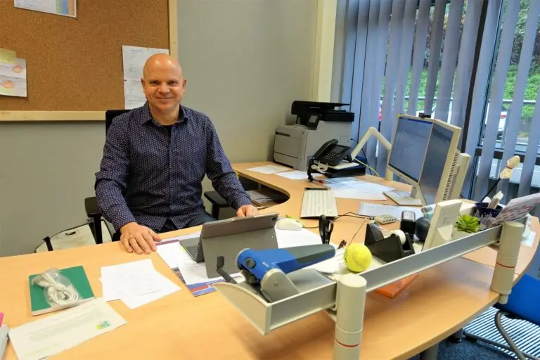 Mann im blauen Hemd sitzt lächelnd an einem Schreibtisch mit Unterlagen im Büro