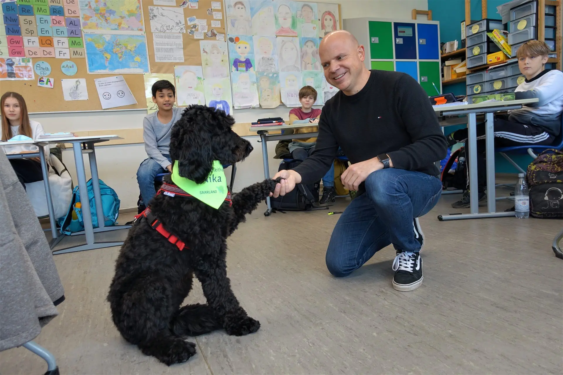 Mann hockt im Klassenraum vor dem schwarzen Schulhund, Schüler im Hintergrund
