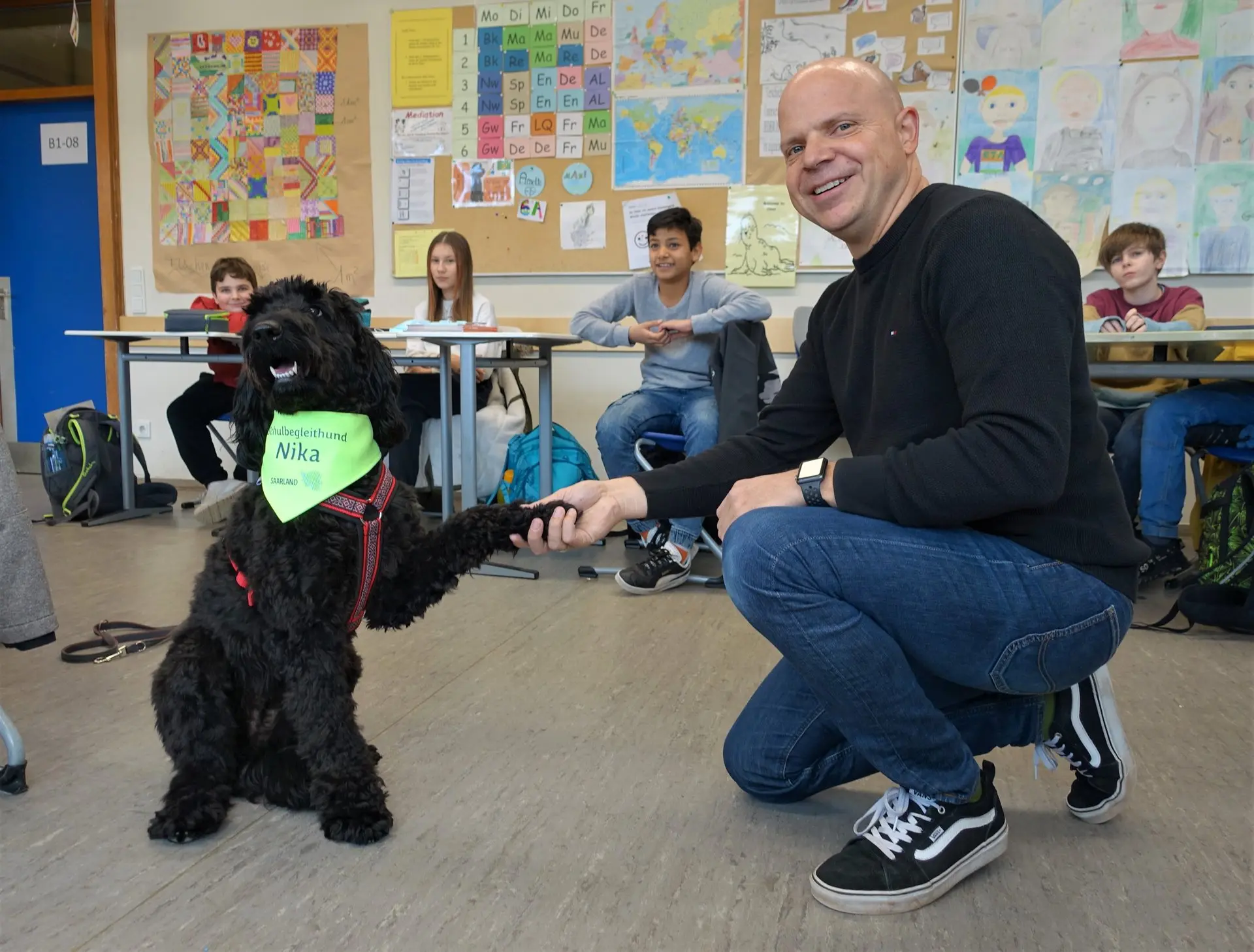 Schwarzer Schulhund mit grünem Halstuch gibt einem hockenden Mann Pfote im Klassenraum