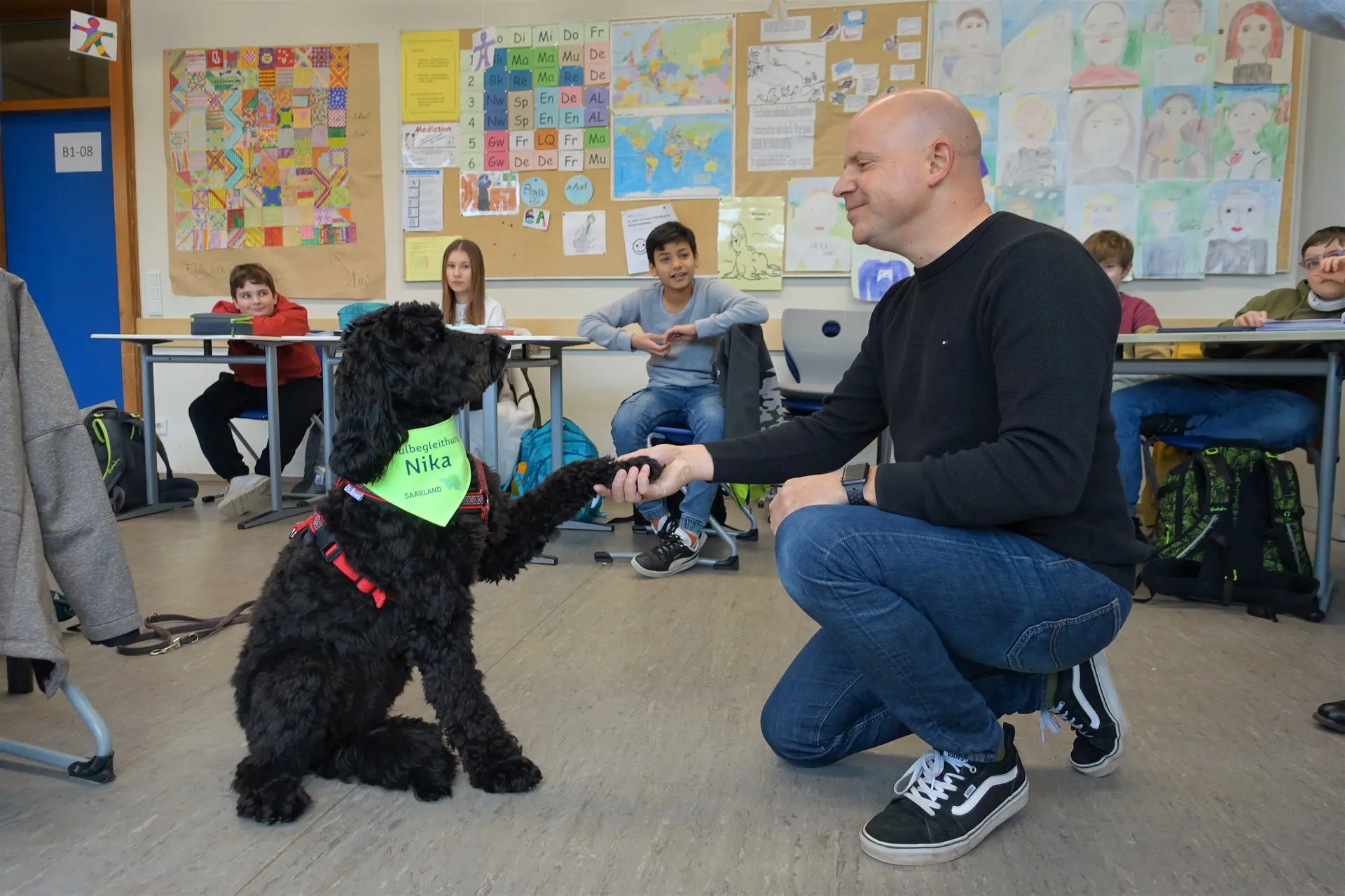 Hund mit grünem Halstuch gibt einem Mann im Klassenraum Pfote, Schüler schauen zu