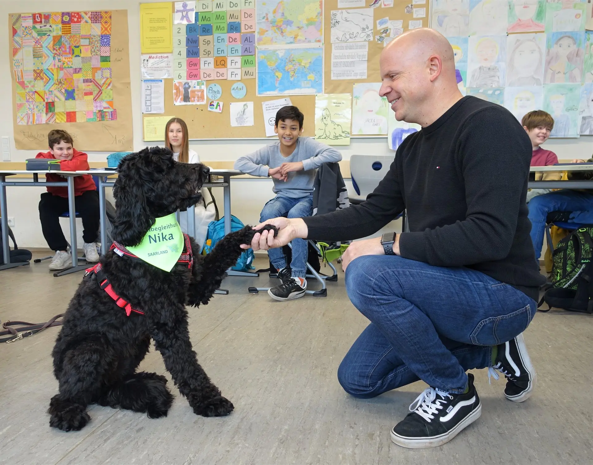 Mann hockt im Klassenraum und hält die Pfote des schwarzen Schulhunds