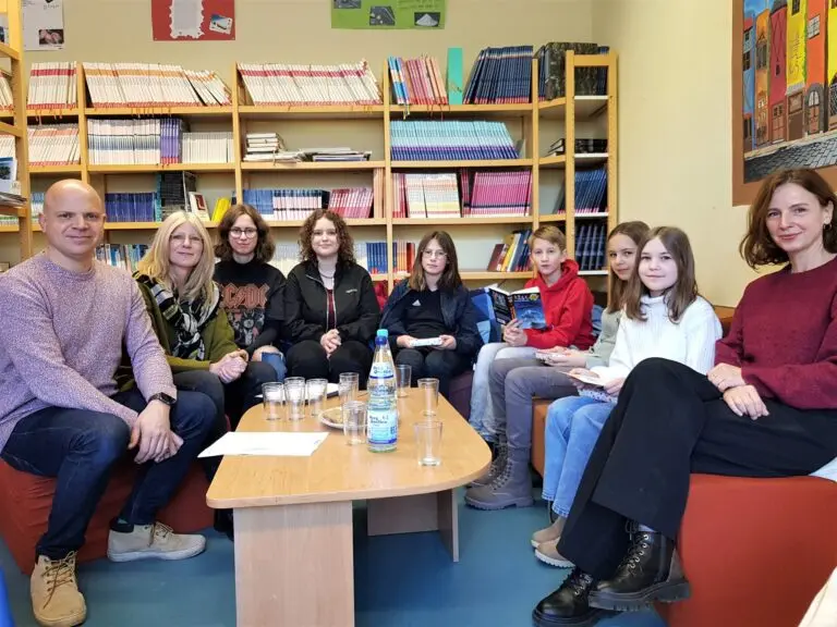Schülerinnen und Erwachsene sitzen im Kreis in einer Schulbibliothek vor Bücherregalen