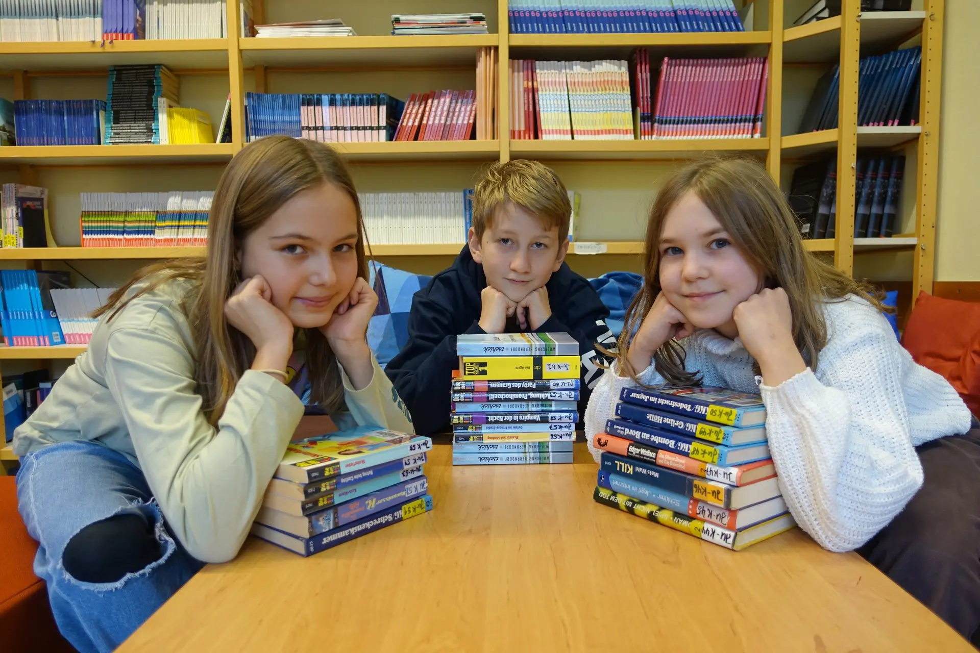 Drei Kinder sitzen an einem Tisch hinter Bücherstapeln in der Bibliothek