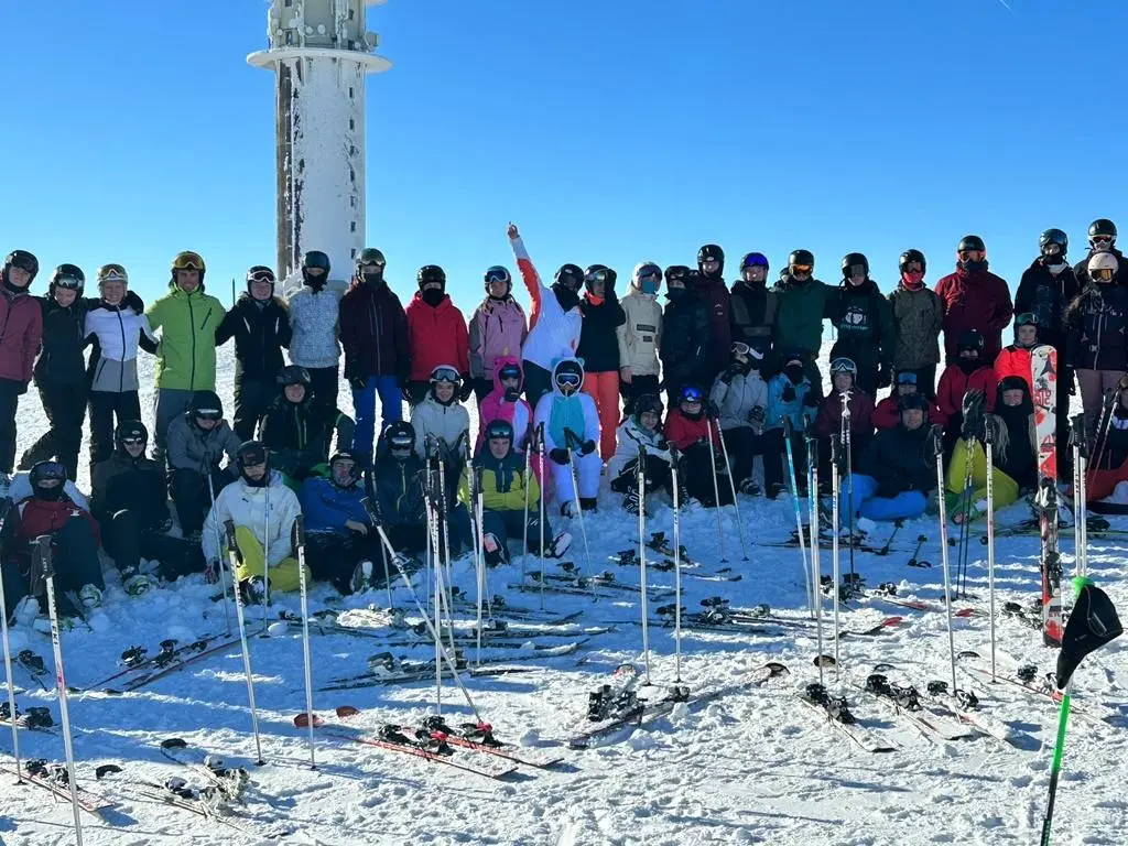 Schülergruppe mit Skiern im Schnee vor einem Turm bei strahlend blauem Himmel