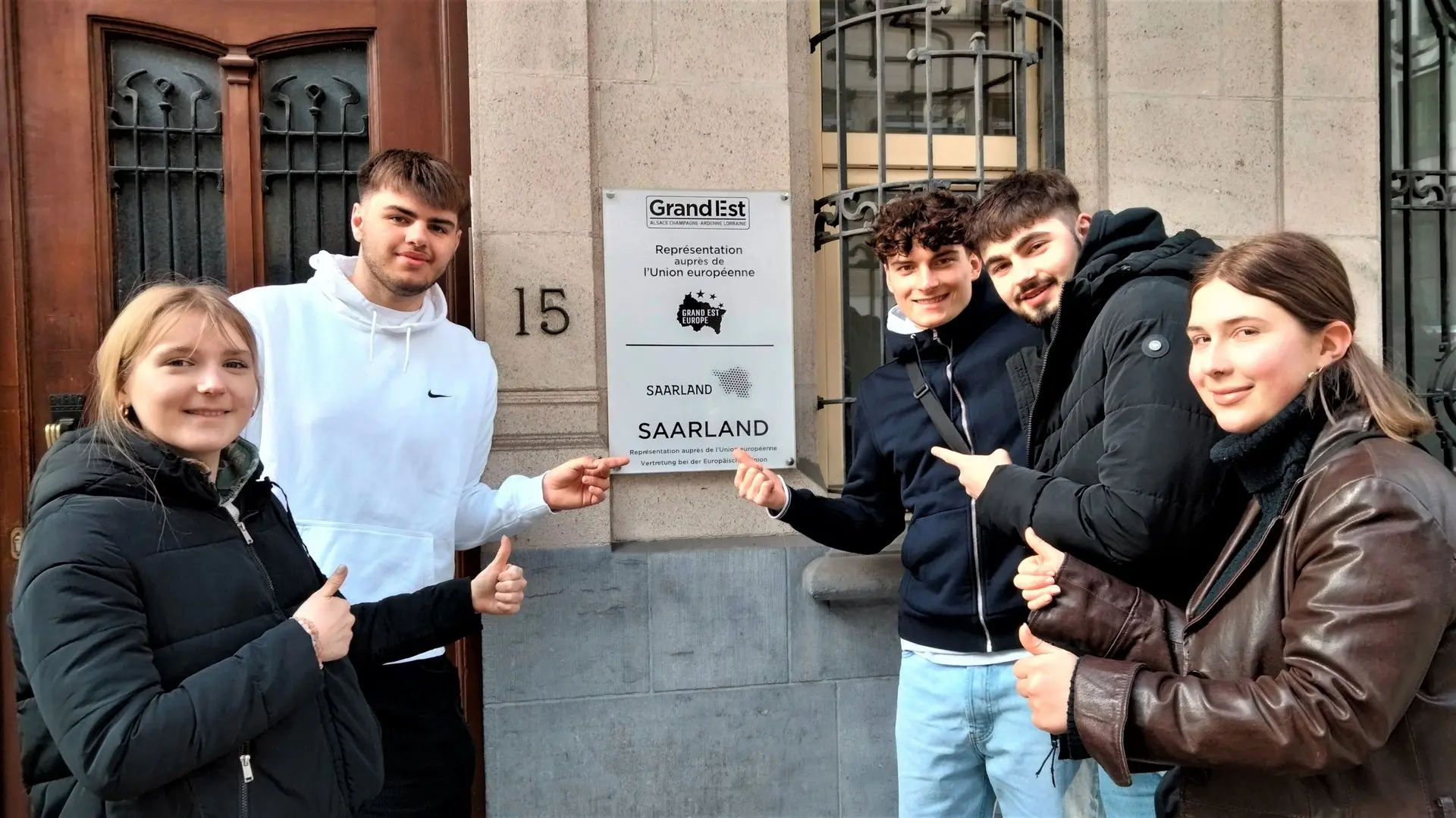 Vier Jugendliche posieren lachend neben einem Hauseingang mit Schild und Hausnummer 15