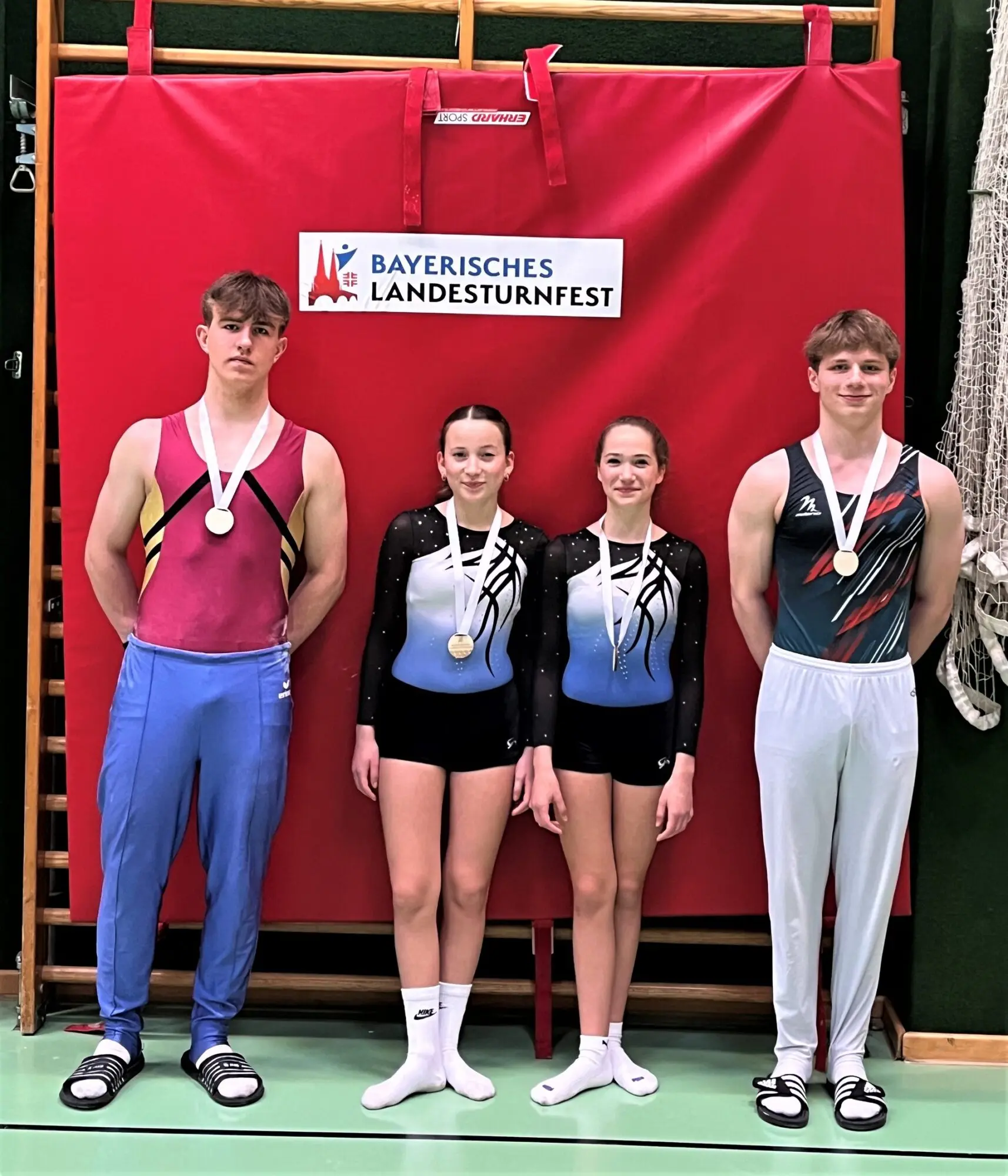 Vier Turnerinnen und Turner mit Medaillen vor einer roten Wand mit Banner des Bayerischen Landesturnfests