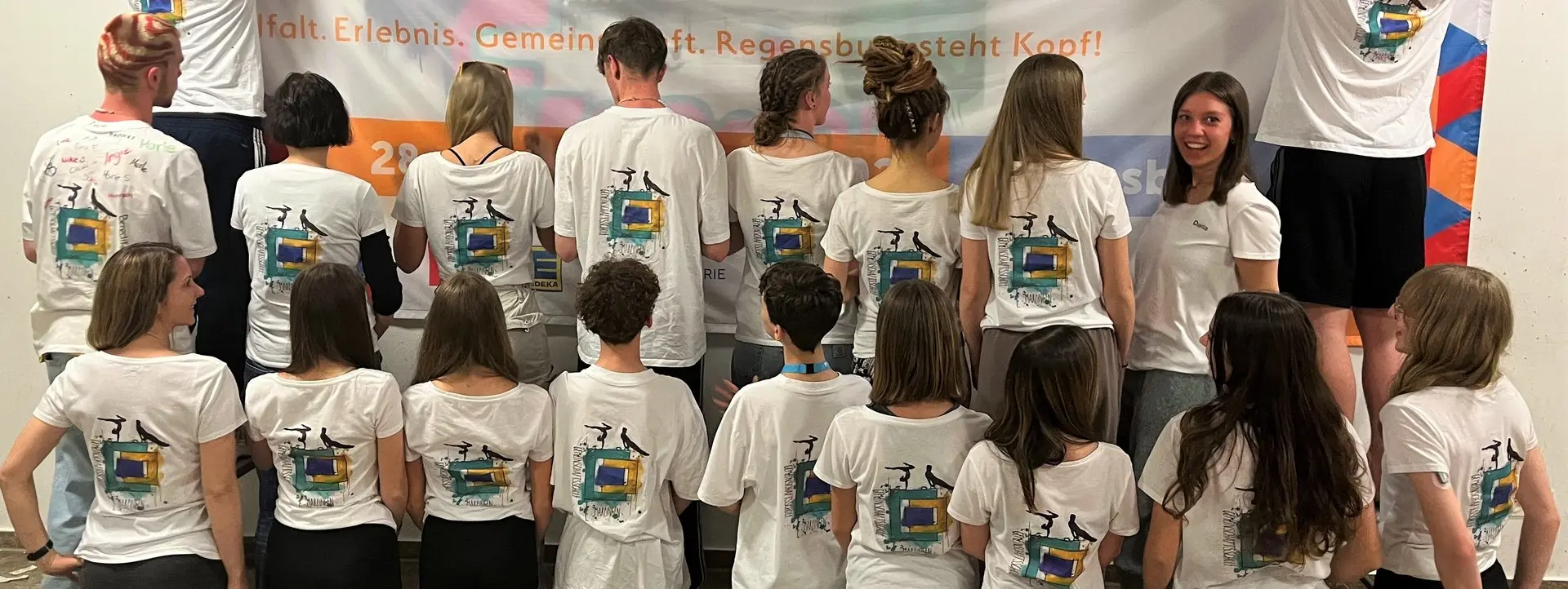 Gruppe Jugendlicher in gleichen weißen T-Shirts, von hinten zu sehen, vor einer Wand