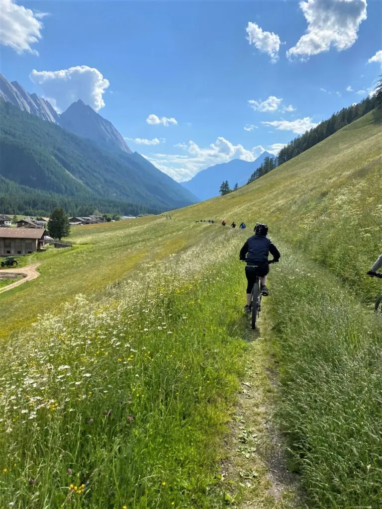 Radfahrer auf einem schmalen Wiesenpfad in einem Hochgebirgstal mit Bergen und blauem Himmel