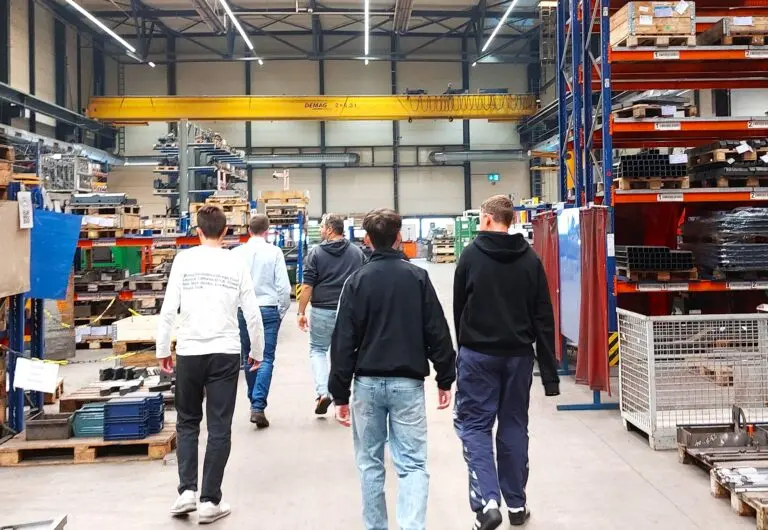 Jugendliche gehen durch eine große Werkshalle mit Hochregalen und Stahlteilen bei einer Betriebsbesichtigung