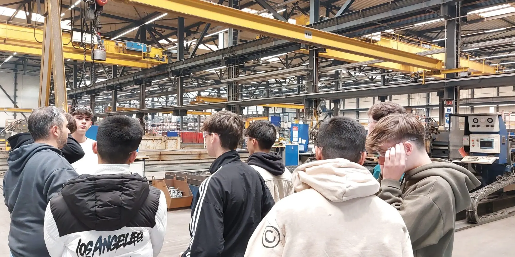 Schülergruppe steht in einer Industriehalle mit gelben Kranbahnen und hört einer Erklärung zu