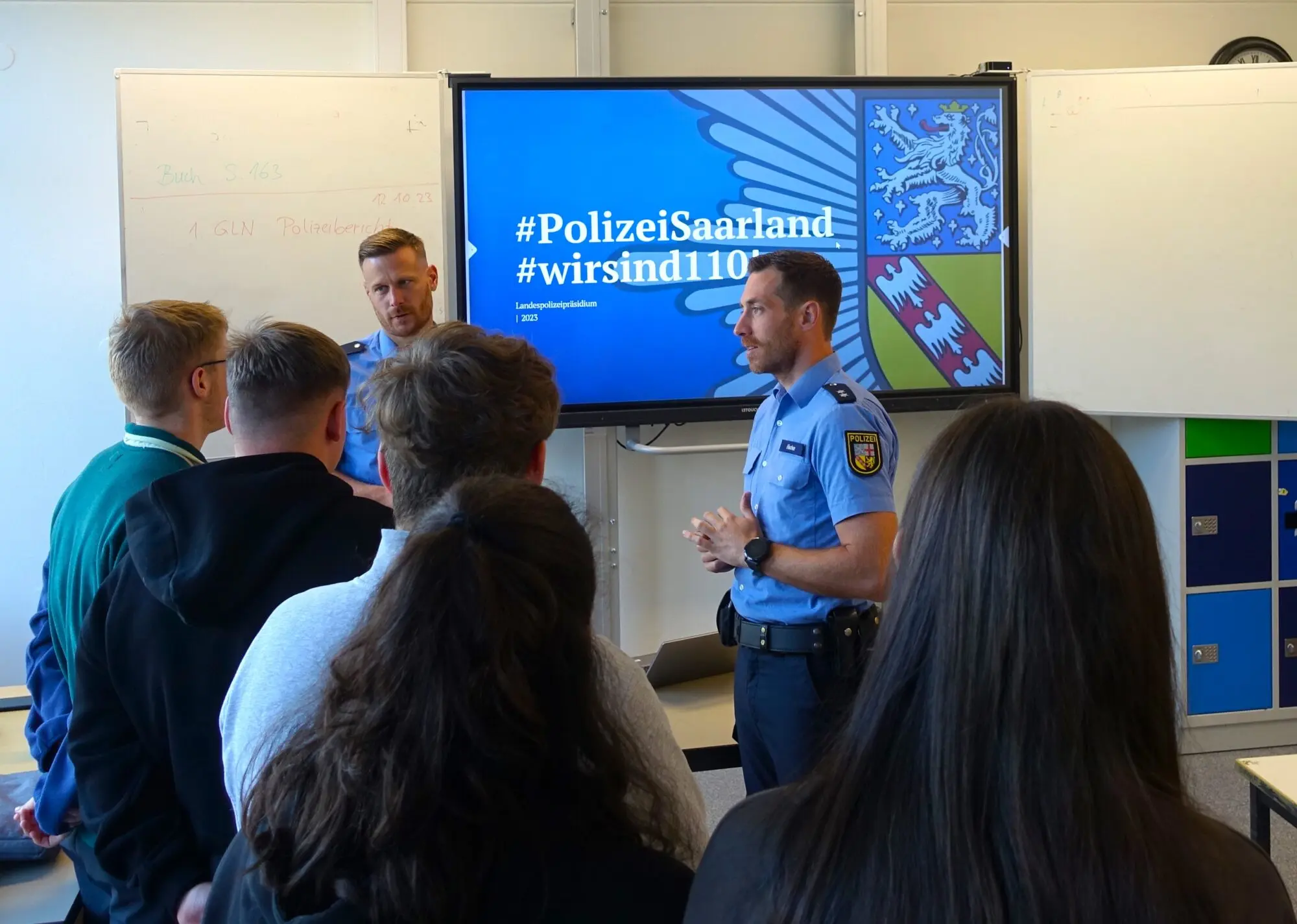 Polizeibeamter stellt Jugendlichen vor einem Bildschirm mit „#PolizeiSaarland“ den Beruf vor