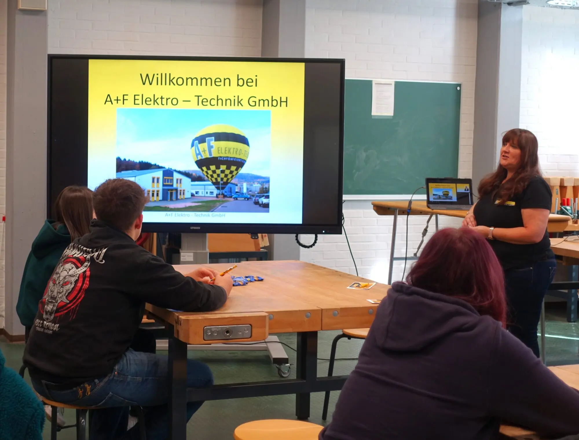 Jugendliche sitzen vor einem Bildschirm mit Willkommensfolie eines Unternehmens, eine Frau präsentiert