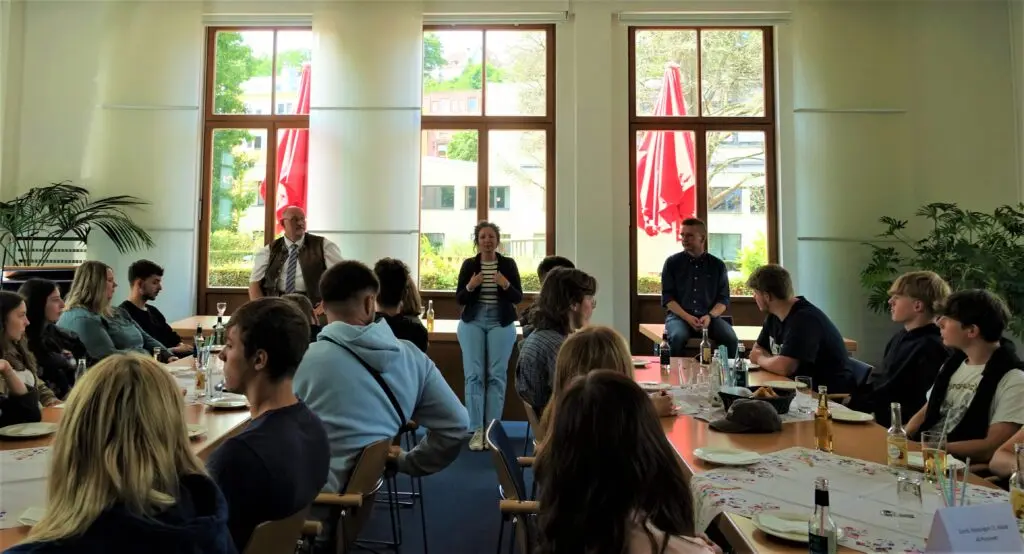 Schülerinnen und Schüler sitzen an gedeckten Tischen in einem hellen Saal