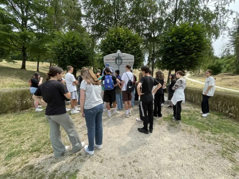 Schülergruppe steht im Kreis an einem Gedenkstein auf einer Wiese, umgeben von Bäumen und Hecken