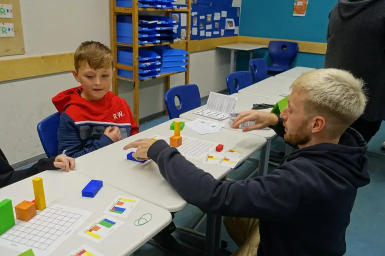 Junge sitzt an einem Tisch mit bunten Bausteinen, ein Mann erklärt ihm eine Aufgabe