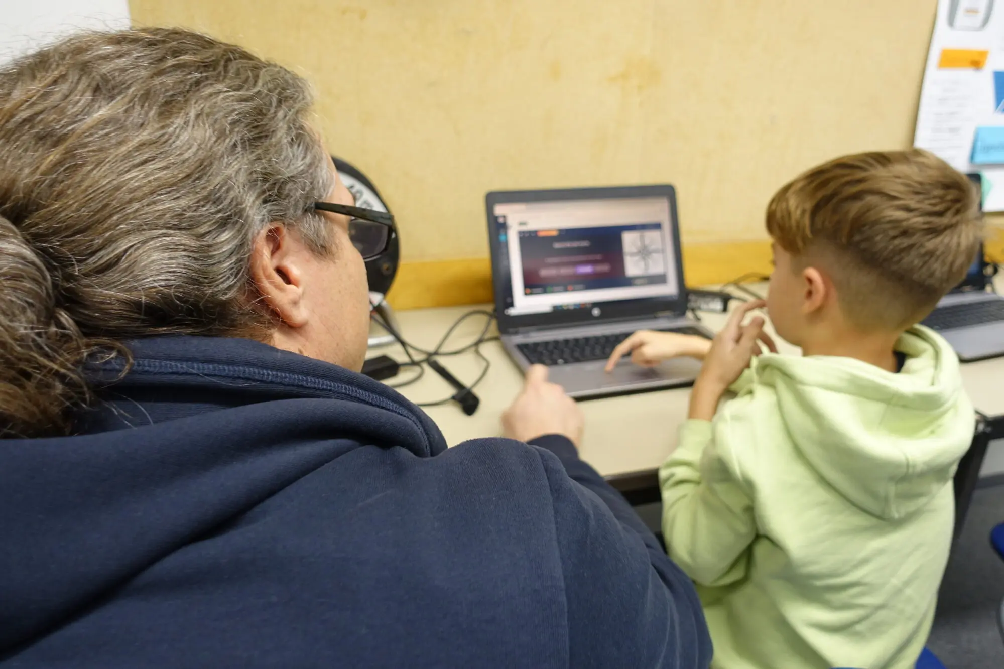 Junge sitzt mit einer Erwachsenen mit Kopfhörern an einem Laptop im Klassenraum