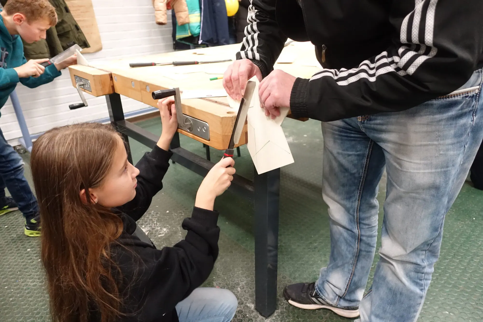Mädchen arbeitet an einer Werkbank mit einem Stück Holz, ein Schüler hilft