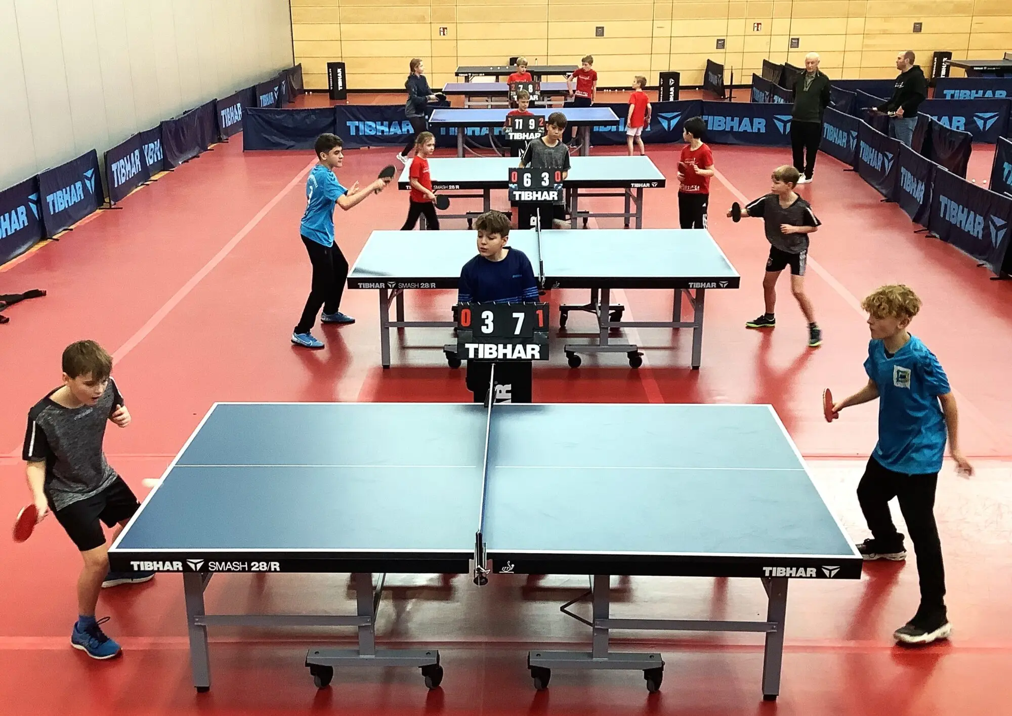 Jungen spielen Tischtennis an mehreren Platten in einer Halle mit rotem Boden