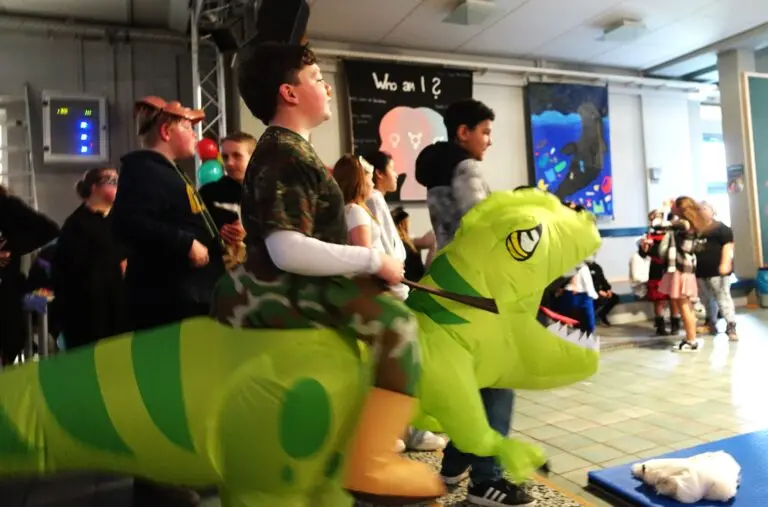 Verkleidete Kinder feiern Fasching in der Aula, vorne ein großer grüner Plüschdrache