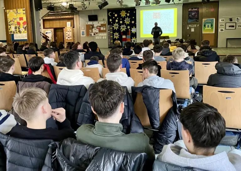 Schülerinnen und Schüler sitzen in einem Saal und verfolgen einen Vortrag auf einer Leinwand
